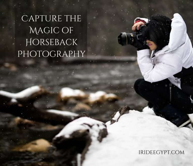 A photographer wearing a white winter jacket and black pants crouches on a snow-covered rock while taking photos in falling snow. Text overlay reads 'CAPTURE THE MAGIC OF HORSEBACK PHOTOGRAPHY' with 'IRIDEEGYPT.COM' in the bottom right corner